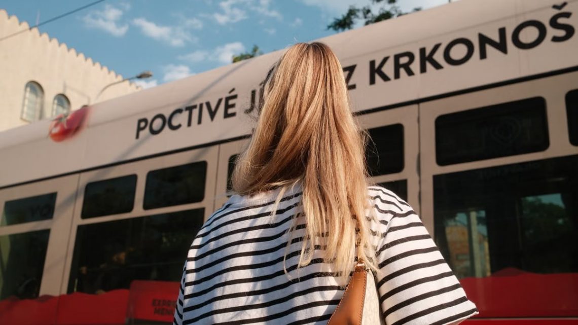 A woman with blonde hair in a striped shirt stands near a city tram on a sunny day.
