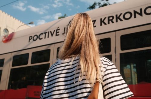 A woman with blonde hair in a striped shirt stands near a city tram on a sunny day.