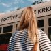 A woman with blonde hair in a striped shirt stands near a city tram on a sunny day.